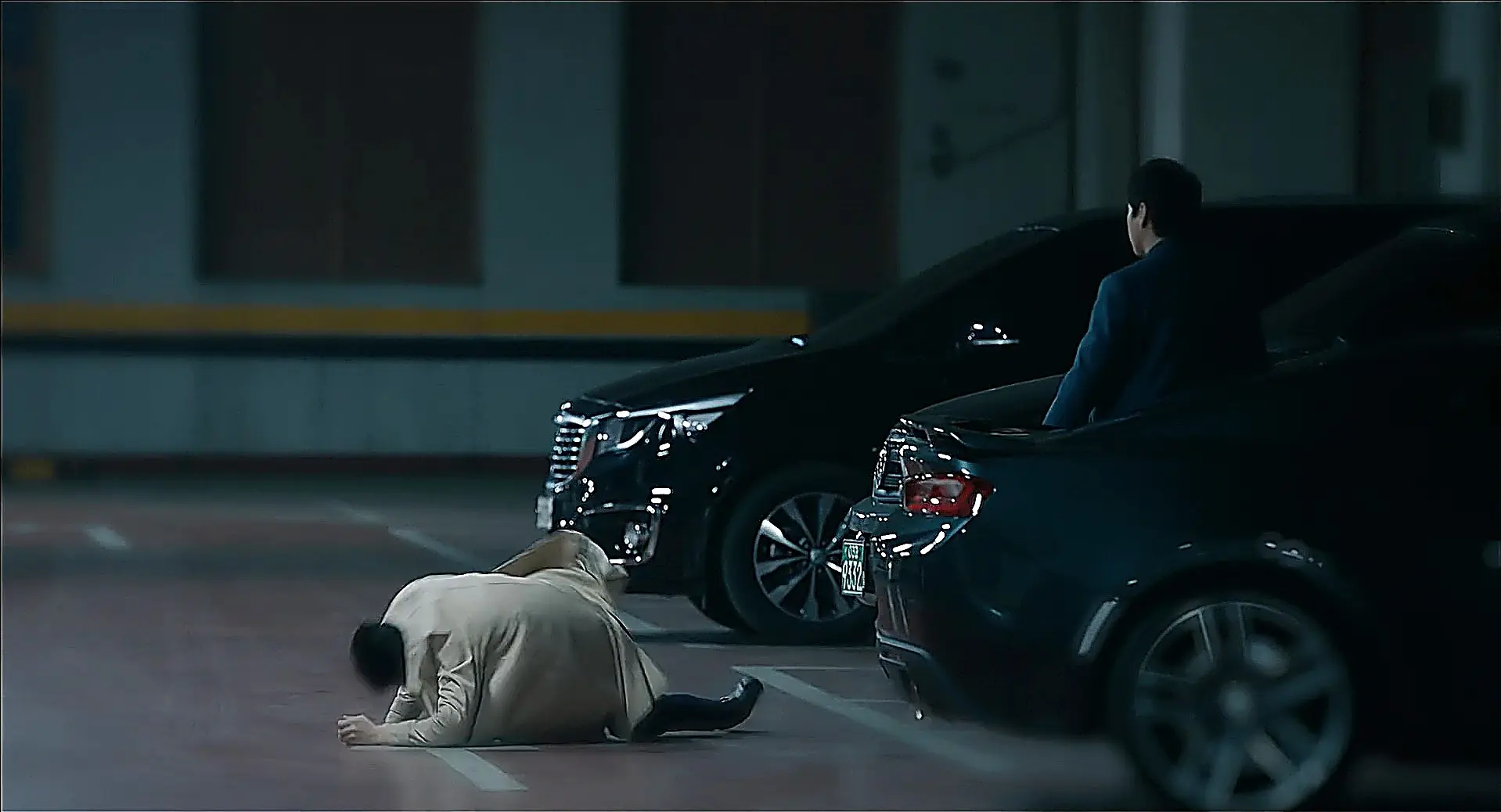A person lying on the ground in an indoor parking structure as a man in a dark suit stands nearby looking down