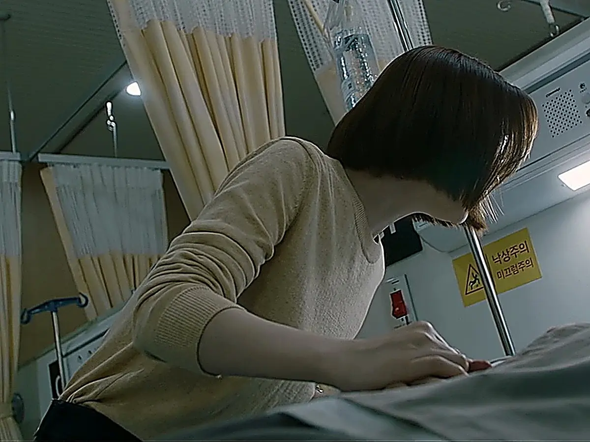 Chae Song-hwa leaning over a hospital bed wearing a beige crewneck long-sleeve knit top