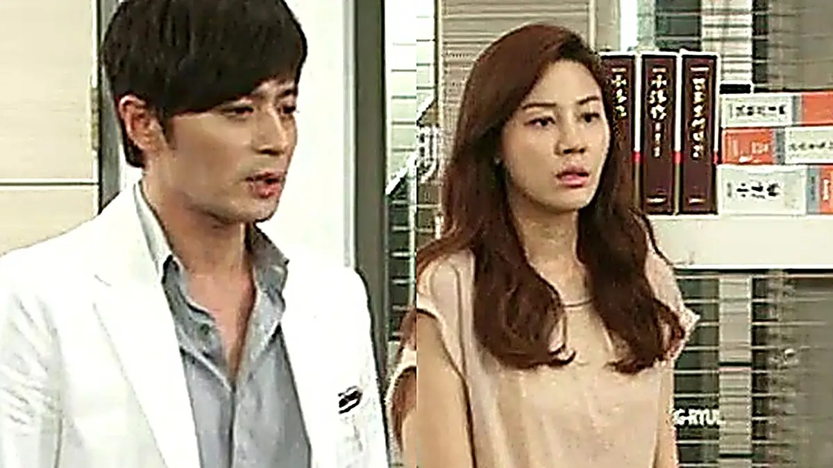 A man in a white jacket over a grey button-up shirt standing beside a woman with wavy brown hair in a light-colored top before a bookshelf