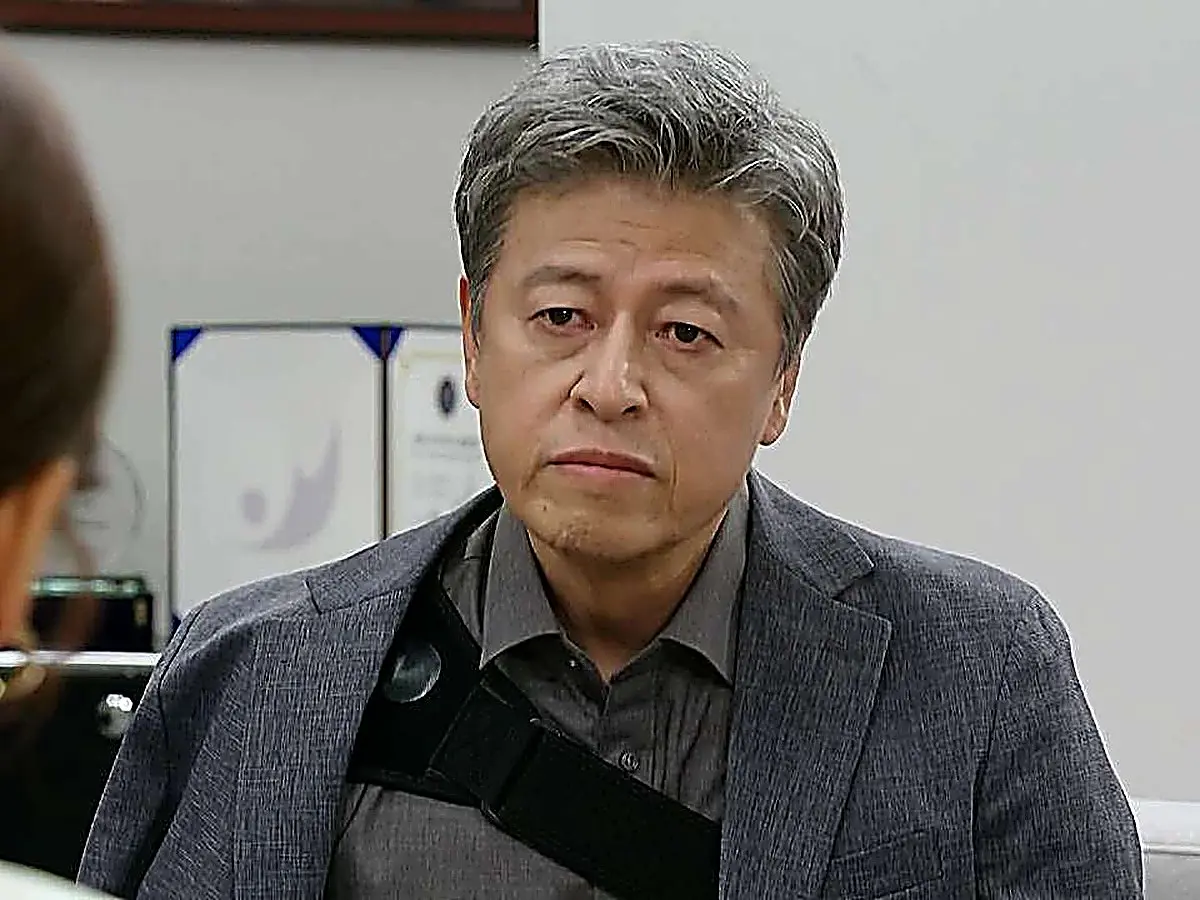 A middle-aged man with grey hair wearing a grey jacket and an arm sling strap looking forward