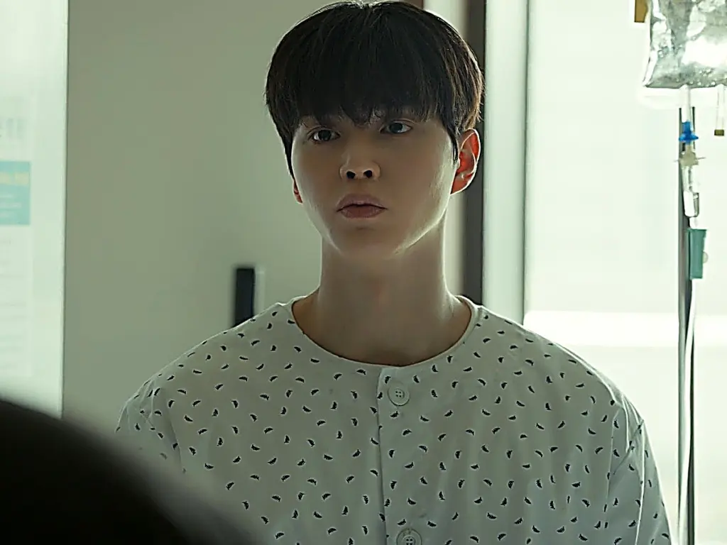 Young man with dark hair in a patterned hospital gown looking straight ahead beside an IV stand