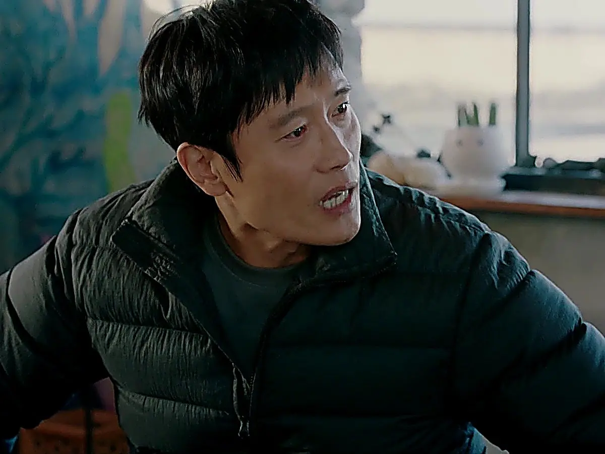 A man with a tense facial expression leaning forward in a dark quilted jacket