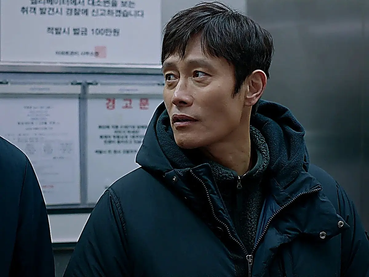 A man in a dark hooded jacket looking sideways inside an elevator with notices on the wall