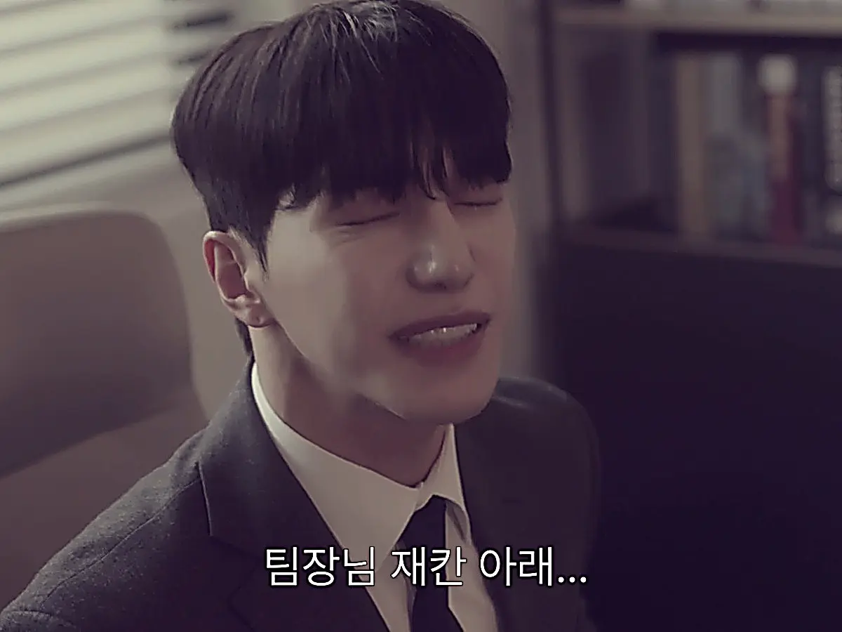Lee Seon-woo sitting in the team lead chair wearing a charcoal tailored suit jacket and tie