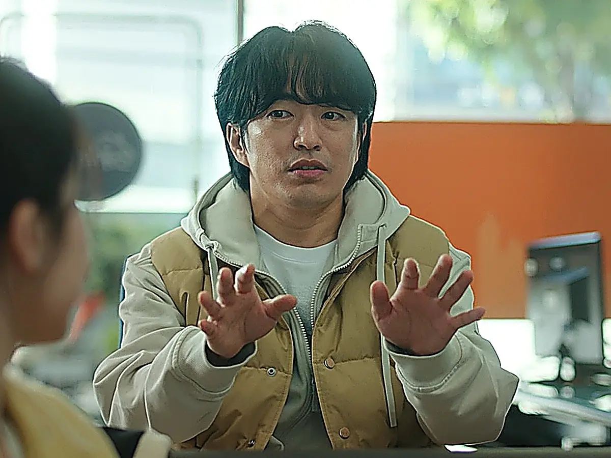 A man wearing a hooded sweatshirt and a tan vest holding up both hands while talking