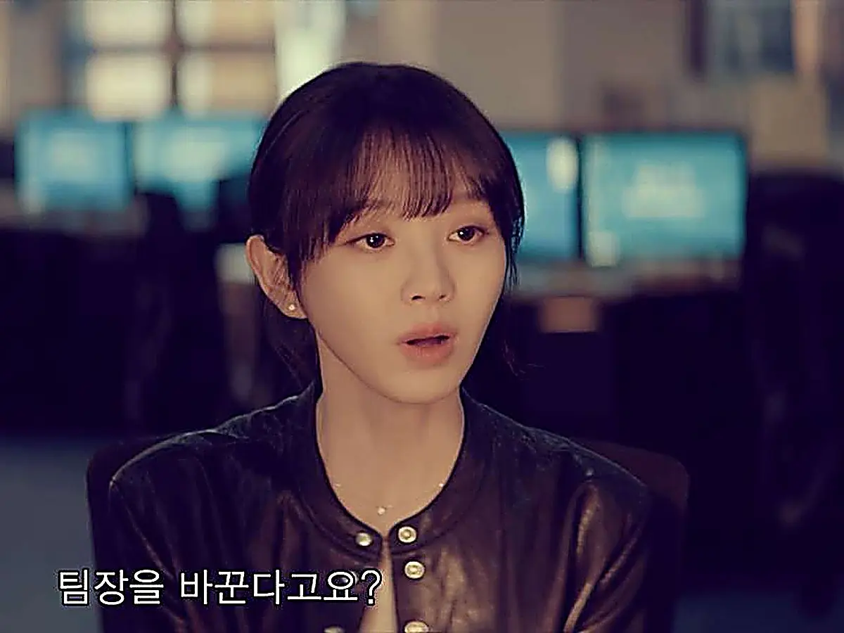 A woman with dark bangs in a black jacket looking surprised in front of computer monitors
