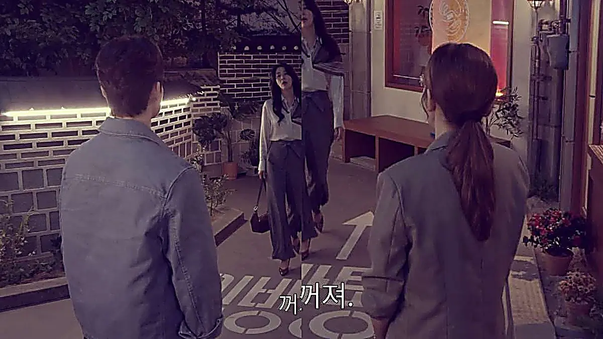 A woman with wavy dark hair walking toward two people viewed from behind along an alleyway