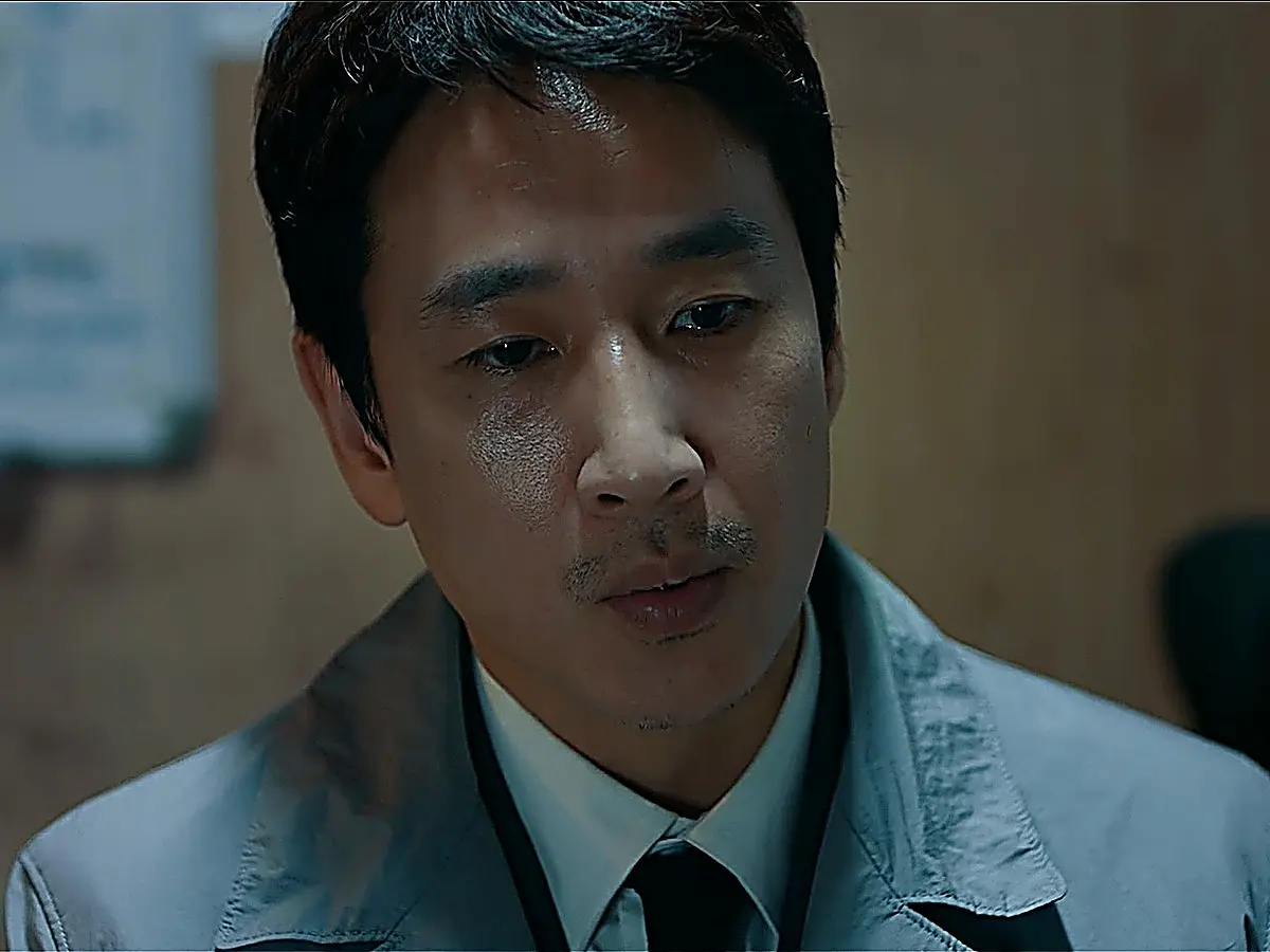 A man in a grey collared coat looking forward with a gentle, attentive gaze