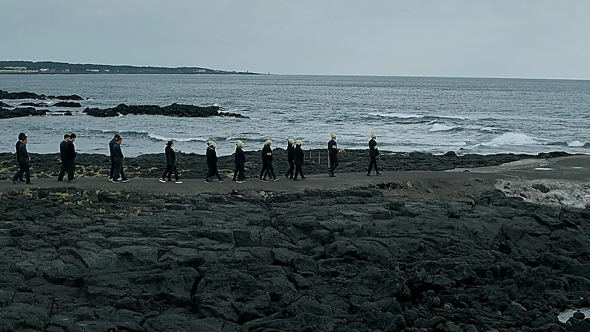 A funeral procession of mourners dressed in black walking along a rocky basalt coastline beside the ocean
