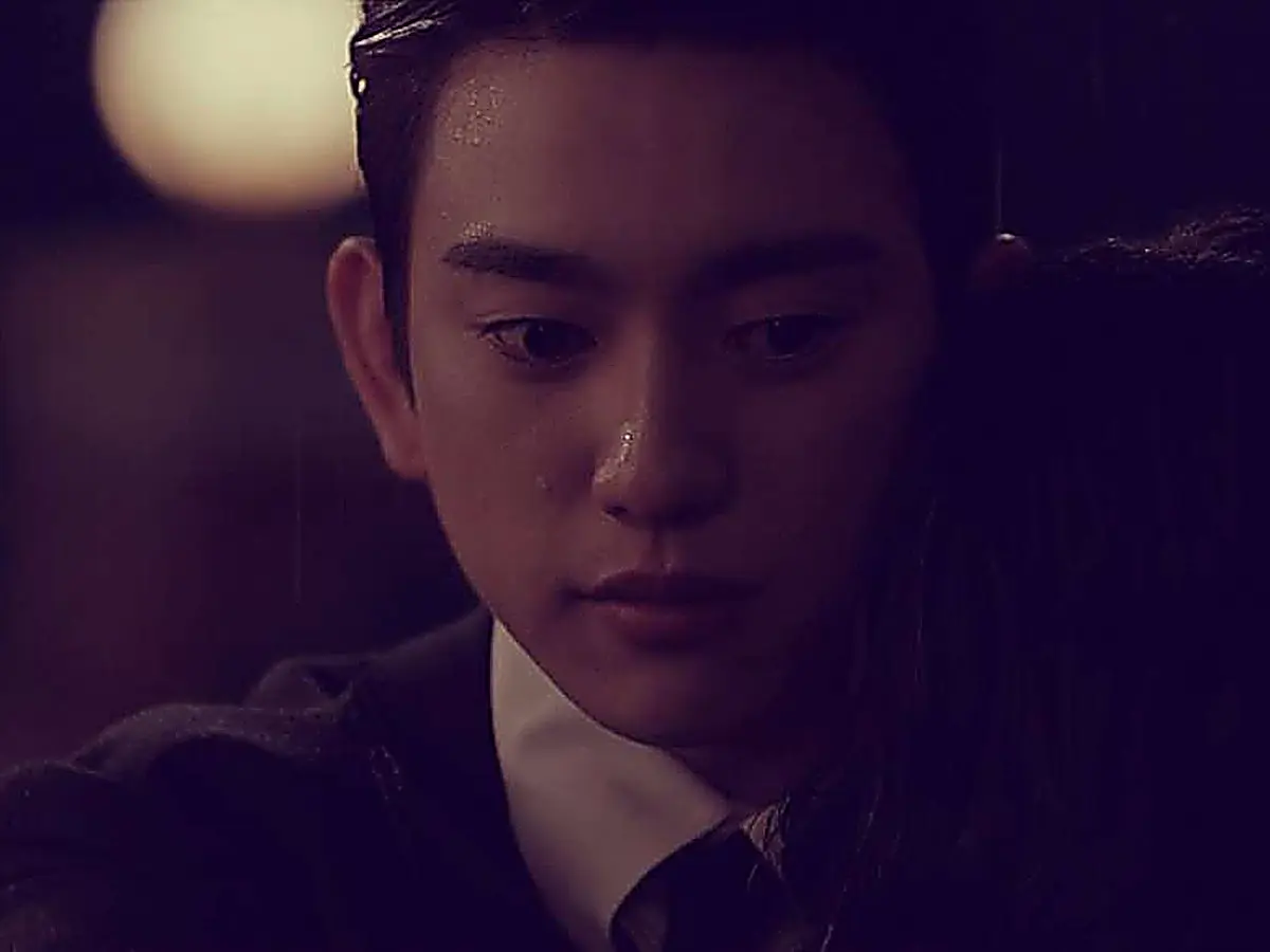 A man with raindrops on his face looking down with a somber expression against a dark background