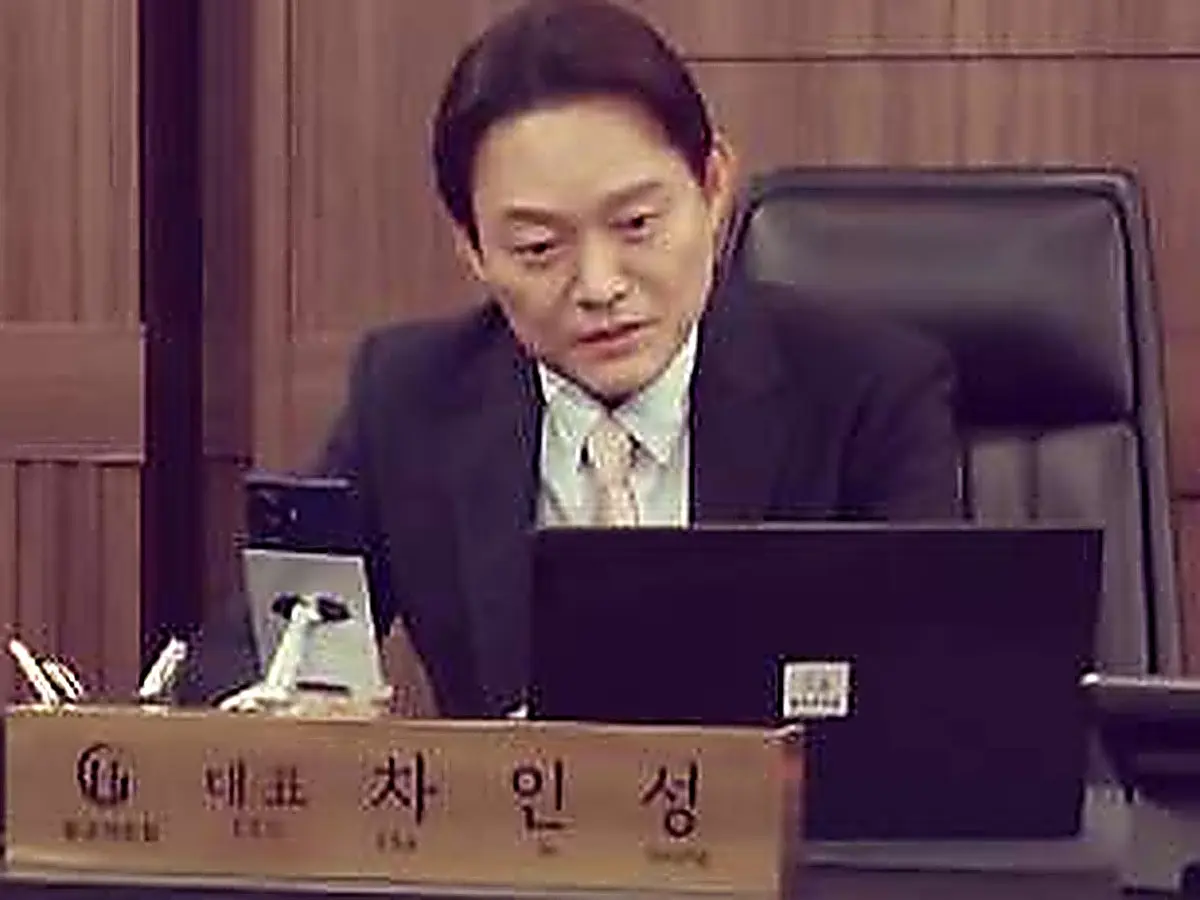 A man in a dark business suit seated at an office desk behind a laptop