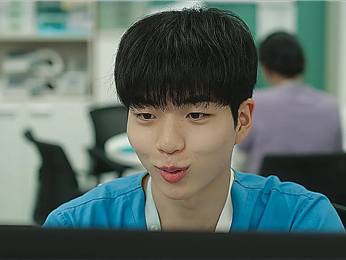 A young man in blue scrubs sitting in front of a screen with a slight smile