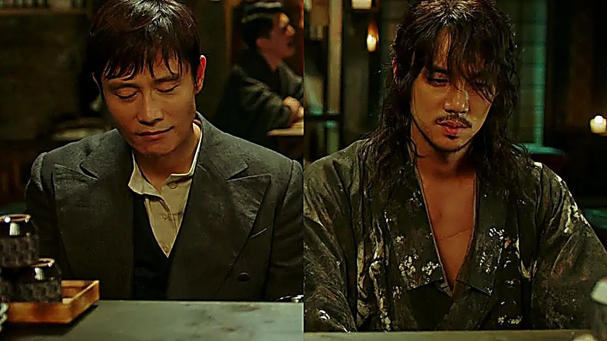 Two men sitting side by side at a wooden counter, one in a dark Western suit and the other in an open dark patterned robe