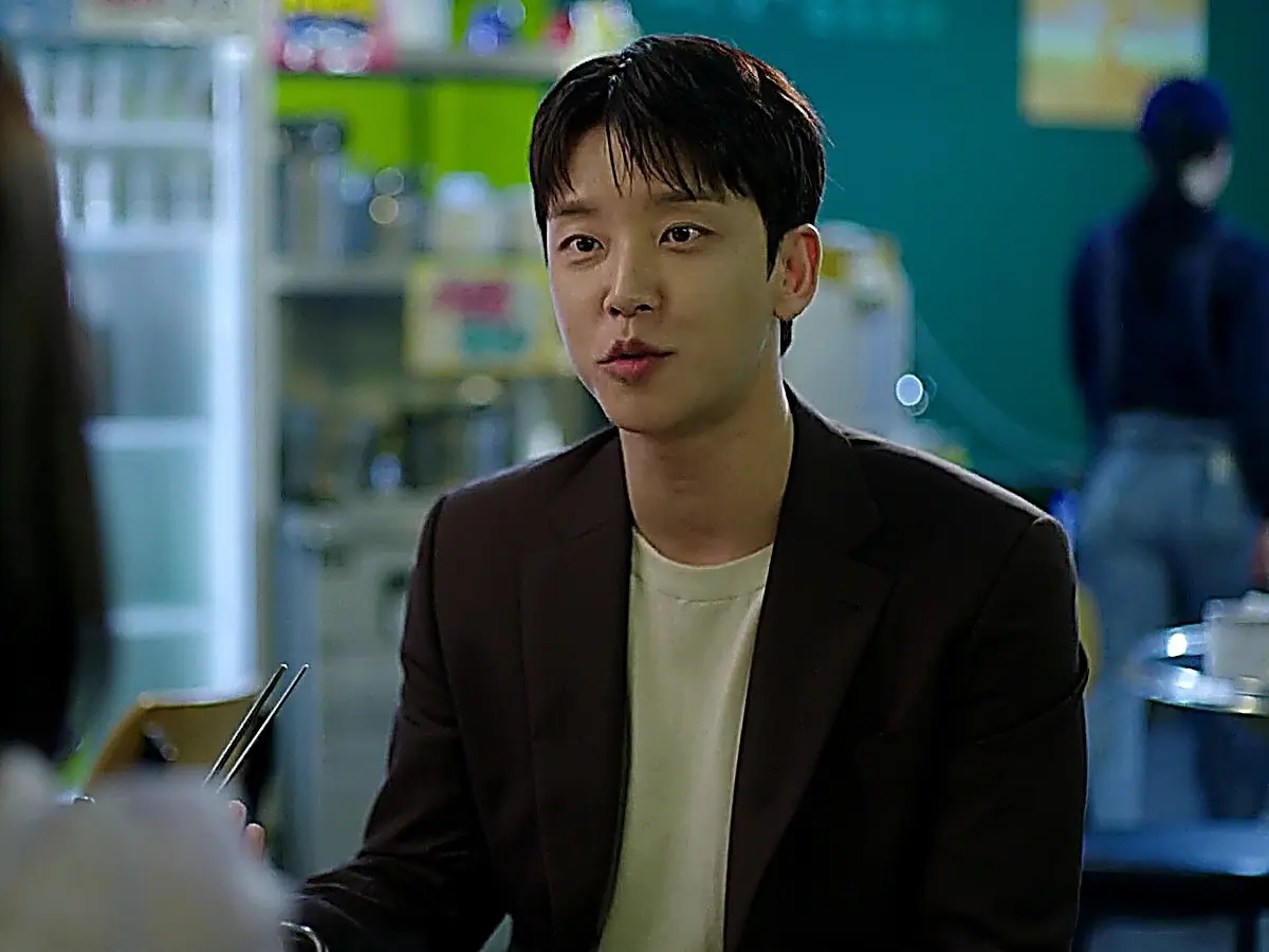 A man in a dark blazer and neutral t-shirt holds chopsticks across a small dining table