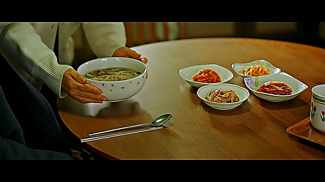 Hands in cream ribbed knit cuffs placing a warm noodle bowl onto a wooden dining table
