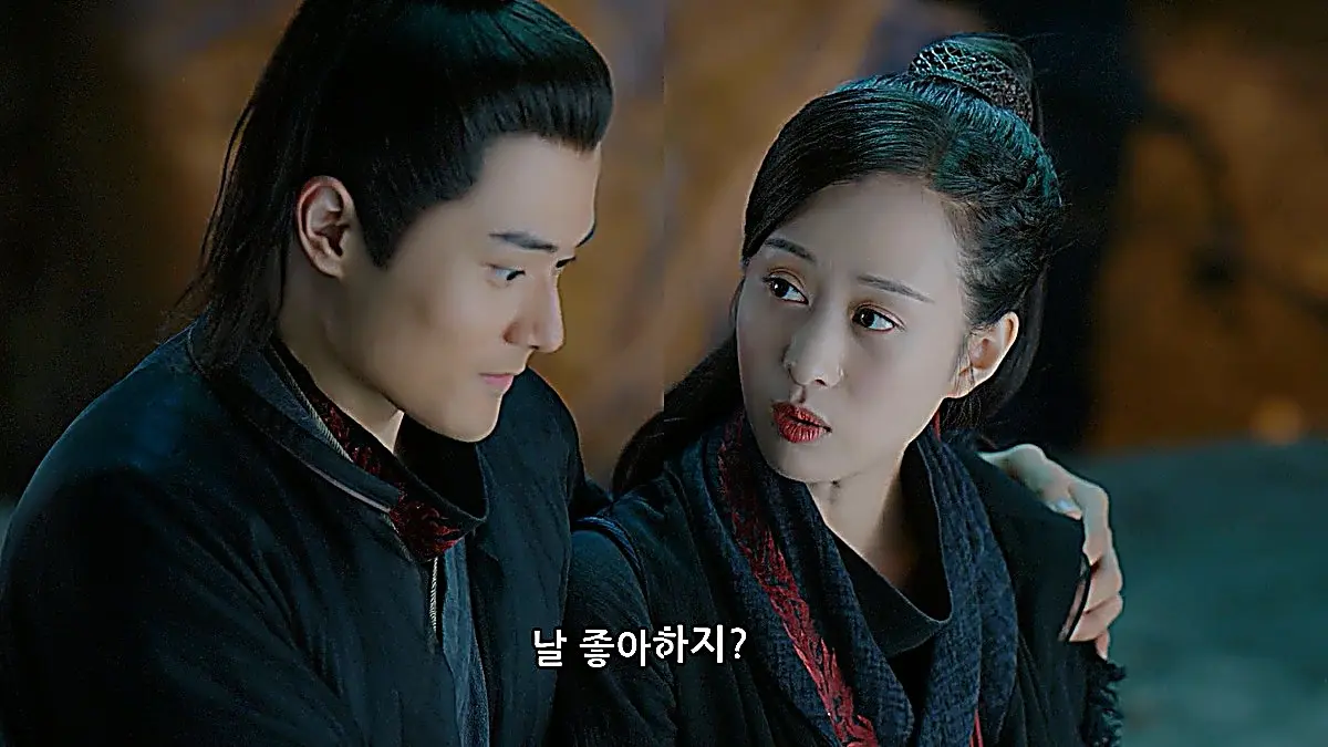 Two people in dark clothing sitting shoulder to shoulder in a dim setting, looking at each other