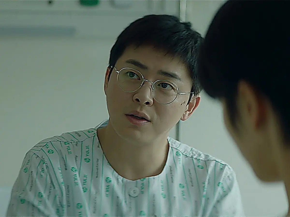 A man with round eyeglasses wearing a patterned hospital patient shirt sitting upright.
