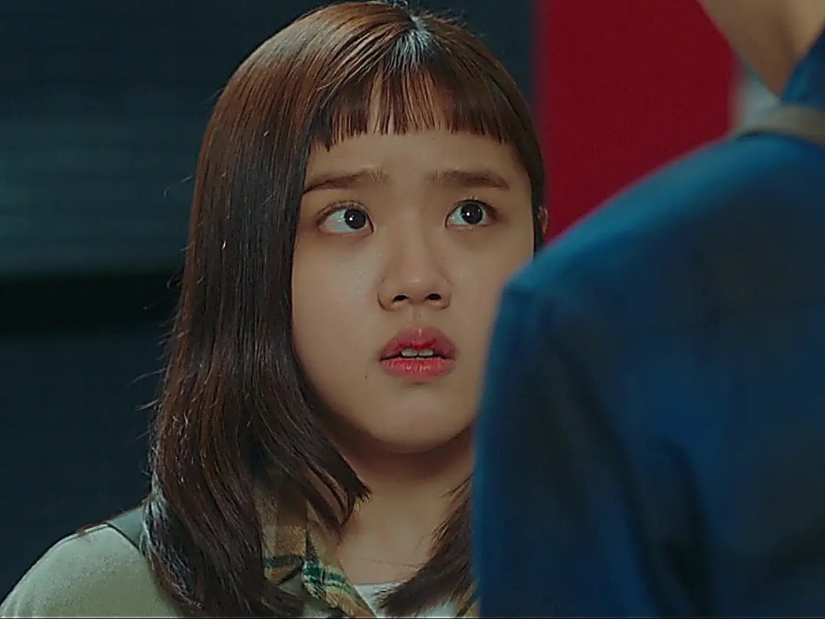 A young woman with straight bangs looking up with wide eyes toward someone wearing a blue shirt