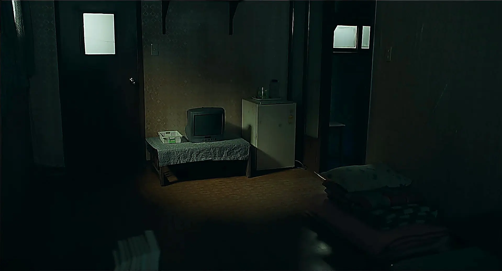 A dimly lit room with a television set, a compact refrigerator, and folded bedding