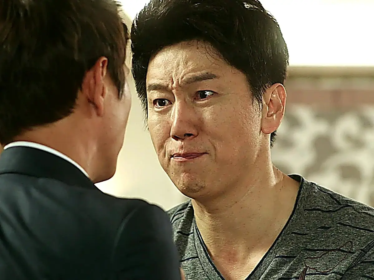 A man in a grey striped t-shirt looking downward with a tearful expression