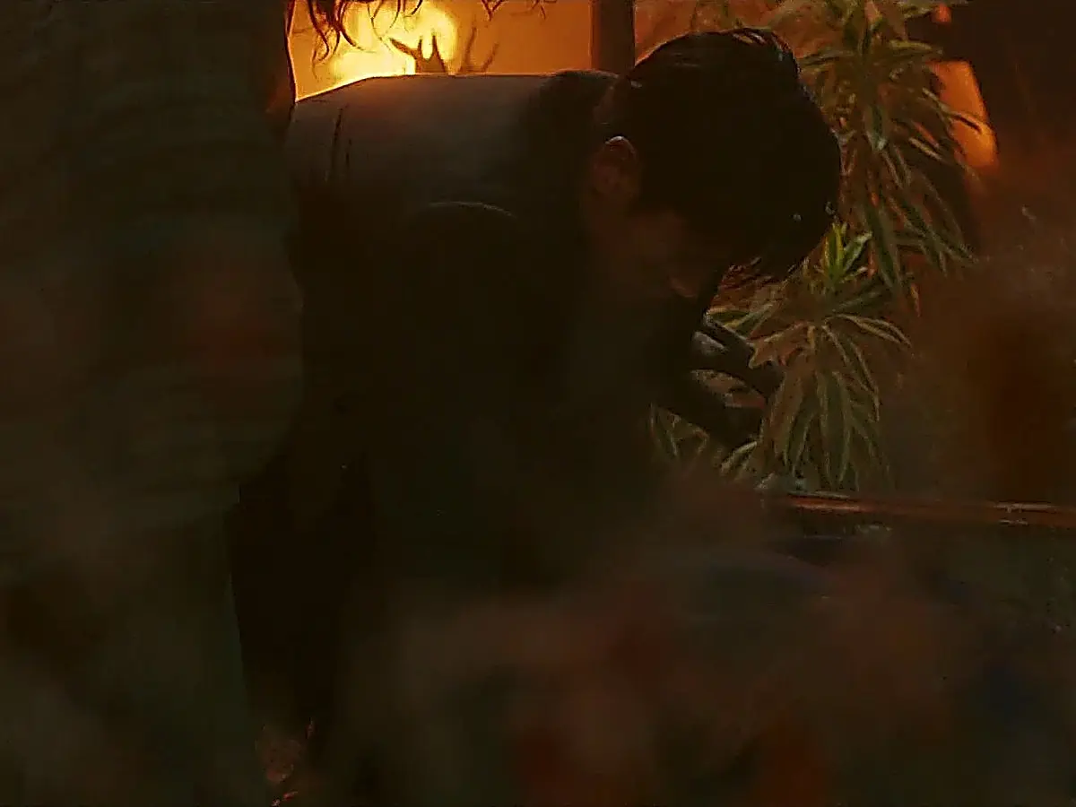 A man in a dark coat moving through smoke and rubble near firelight