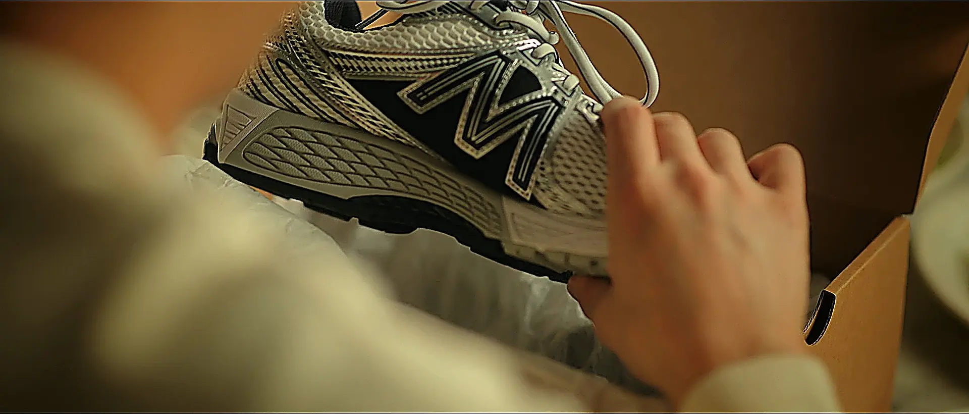 A pair of hands opening a cardboard box containing a silver and black running shoe