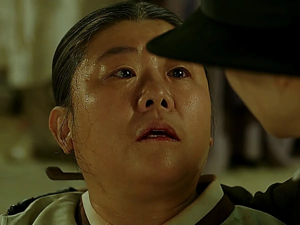 A woman lying down with a pale, strained expression looking up at a person in a black hat