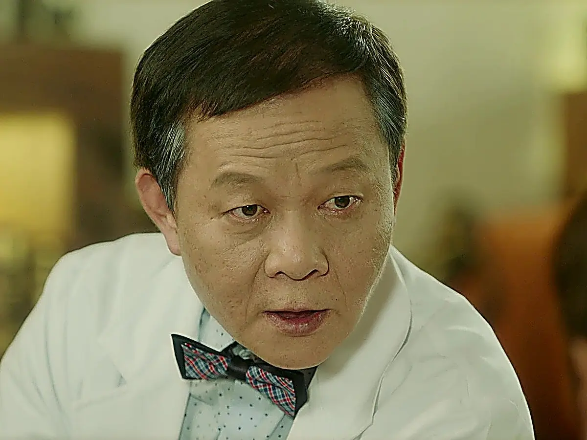Close-up of an older man with short parted hair and glasses wearing a white medical coat and patterned bow tie