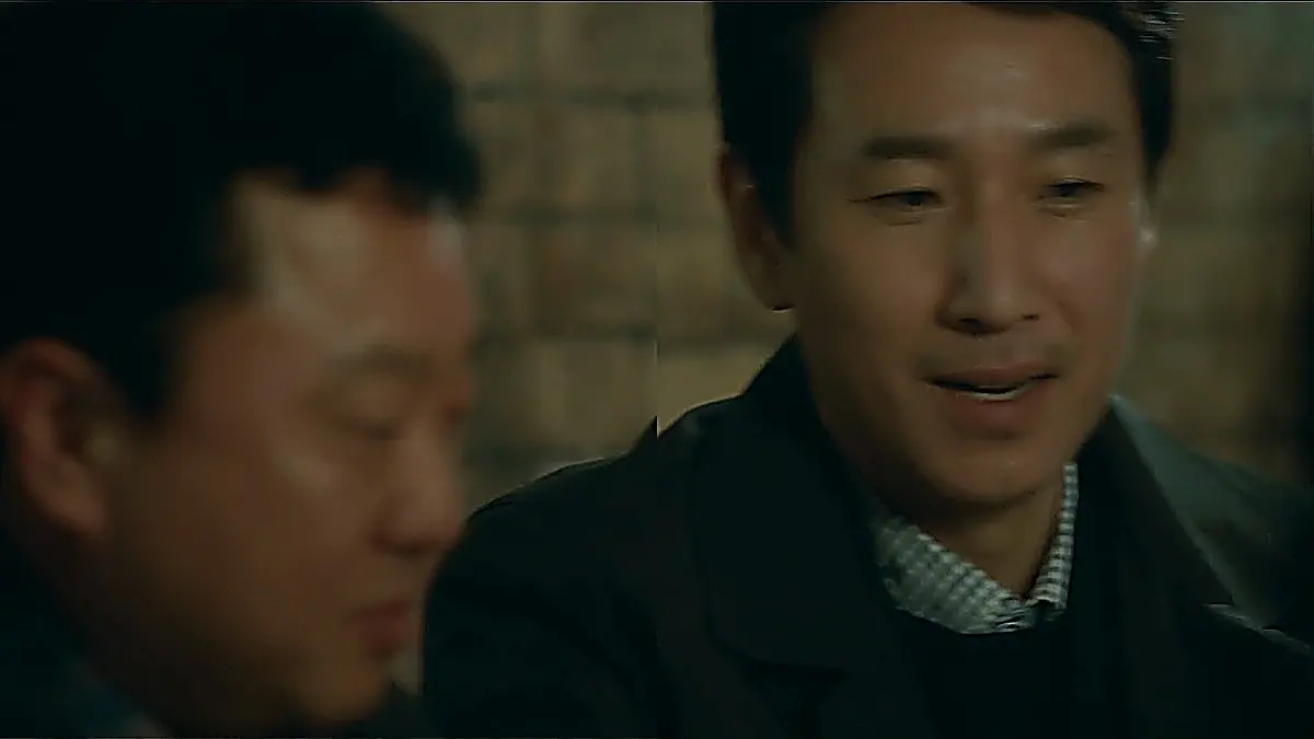 A man smiling while seated beside another man at an indoor gathering