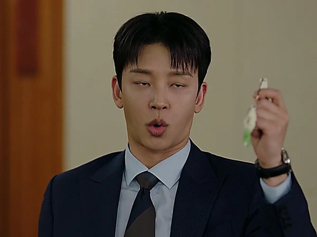 Cha Seok-jin wearing a dark navy suit jacket, light blue collared dress shirt, and necktie while holding up a USB keychain