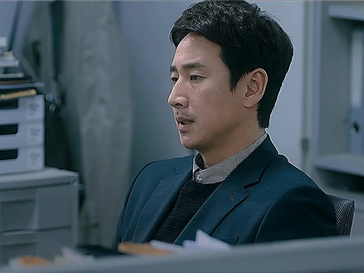A man in a dark suit jacket and collared shirt sitting at an office desk in profile