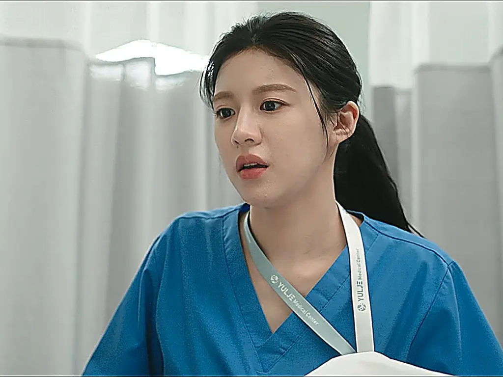 A woman with her hair tied back wearing blue surgical scrubs and a hospital badge lanyard