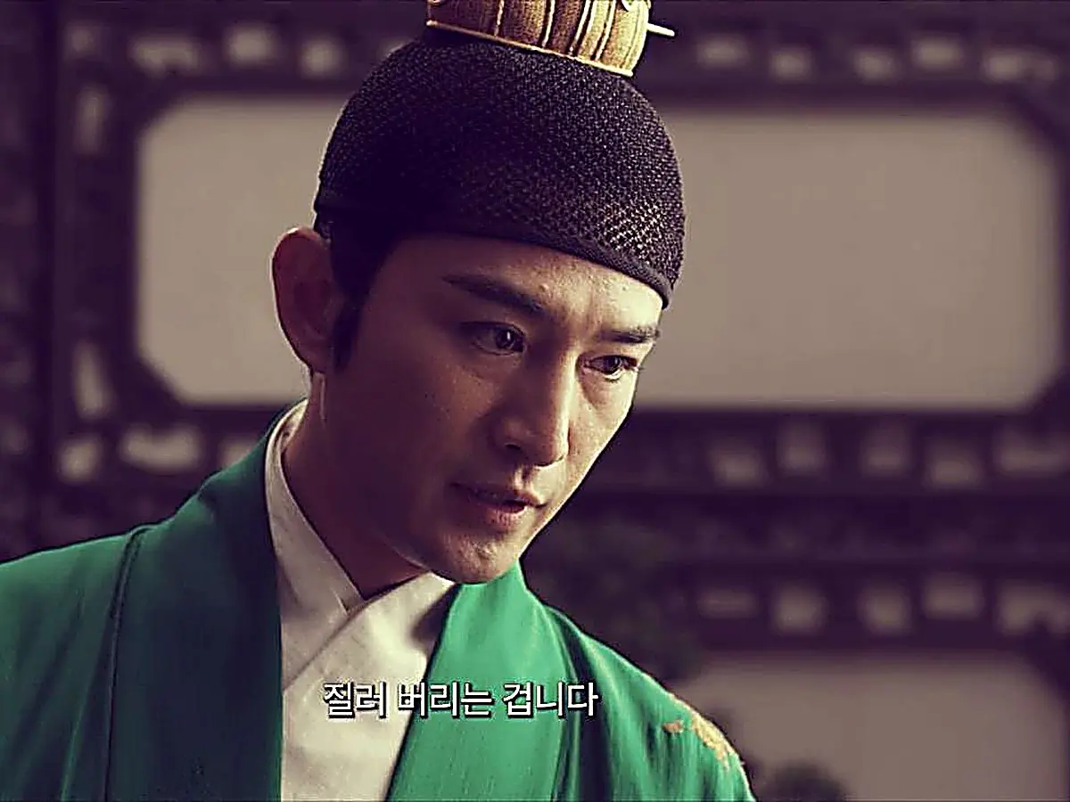 A man wearing a green robe and gold-accented headpiece looking down thoughtfully
