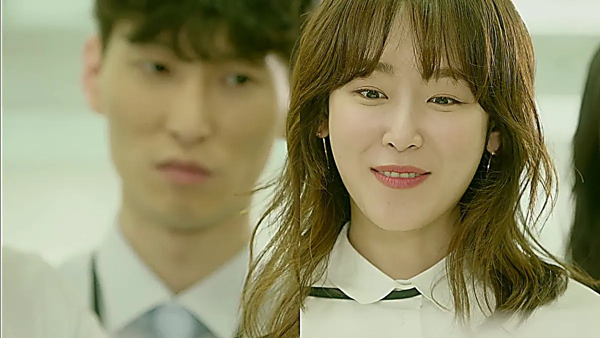 Woman with bangs and wavy hair smiling in a white collared shirt with a blurred man in the background