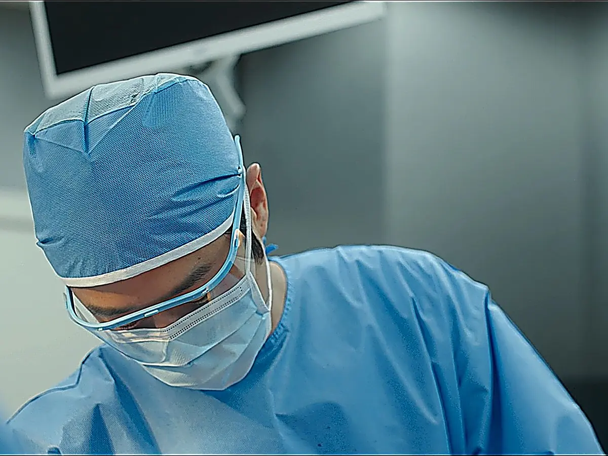 A surgeon wearing a blue cap, protective goggles, and a face mask looking down at an operating table