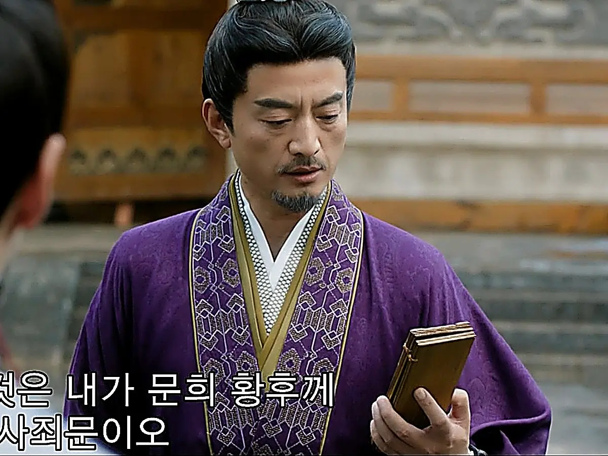 A man with a goatee in a purple patterned robe holding a folded wooden plaque