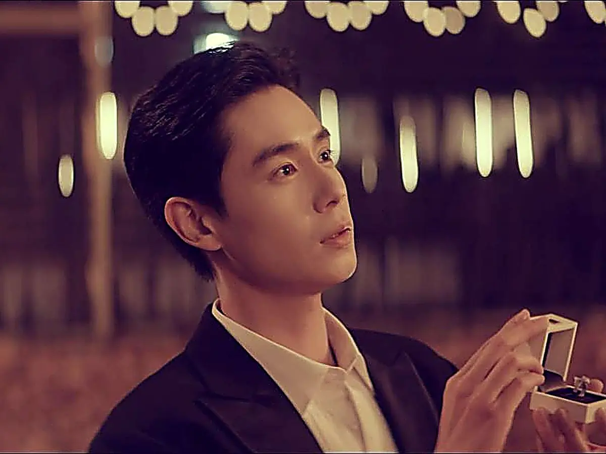 Jiang Yubai holding an open ring box while dressed in a dark tailored suit jacket and white collared shirt