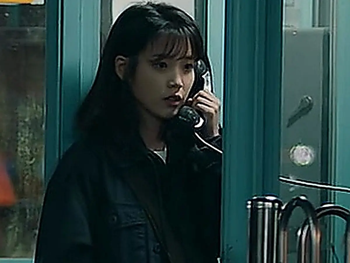 A woman holding a telephone handset inside a teal phone booth