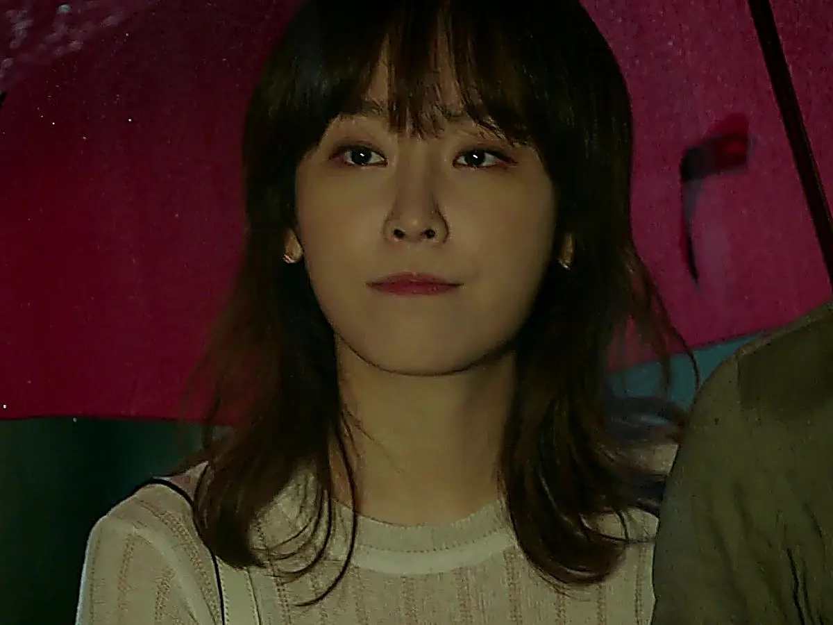A woman with bangs gazes forward under a pink umbrella at night.