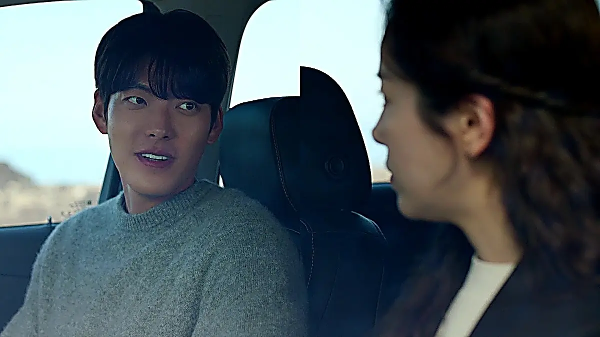 A man in a grey knit sweater speaking from the driver seat of a vehicle while turning toward a passenger