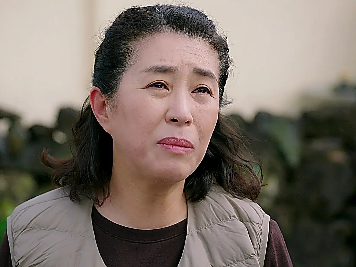 A middle-aged woman with pulled-back dark hair wearing a beige vest looking forward with a tearful expression