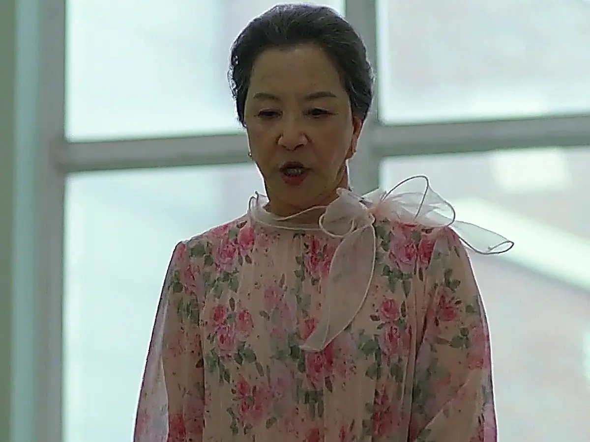 An older woman with grey hair wearing a pink floral sheer blouse stands in a school corridor