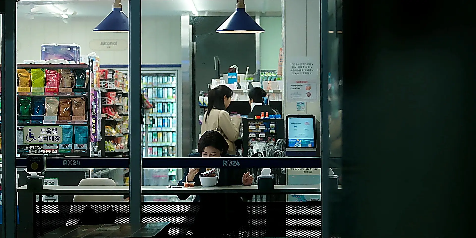 Martina seated at a convenience store counter wearing a tailored jacket over a white blouse