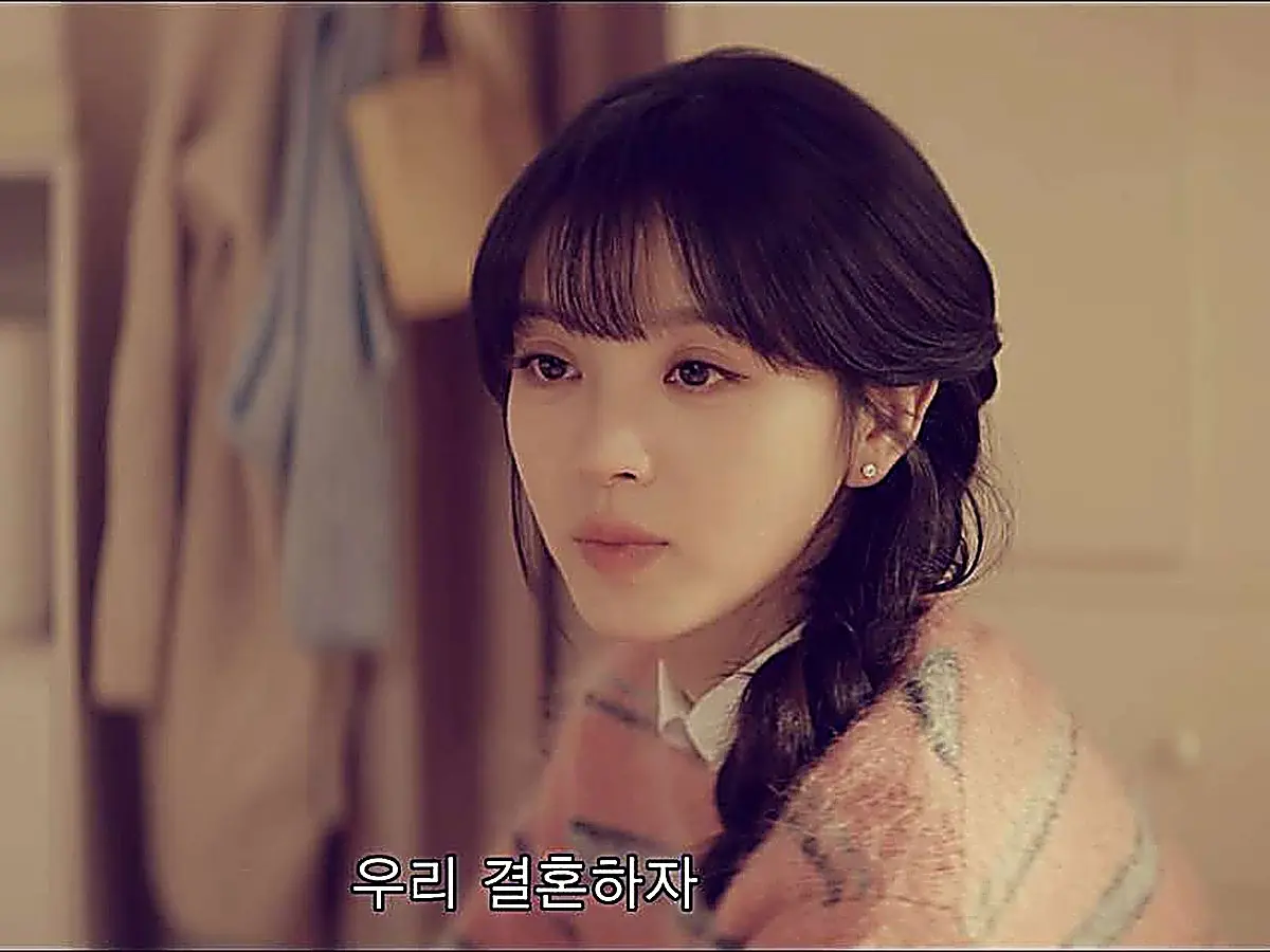 A woman with braided hair wearing a pink knit sweater and collared shirt looking forward