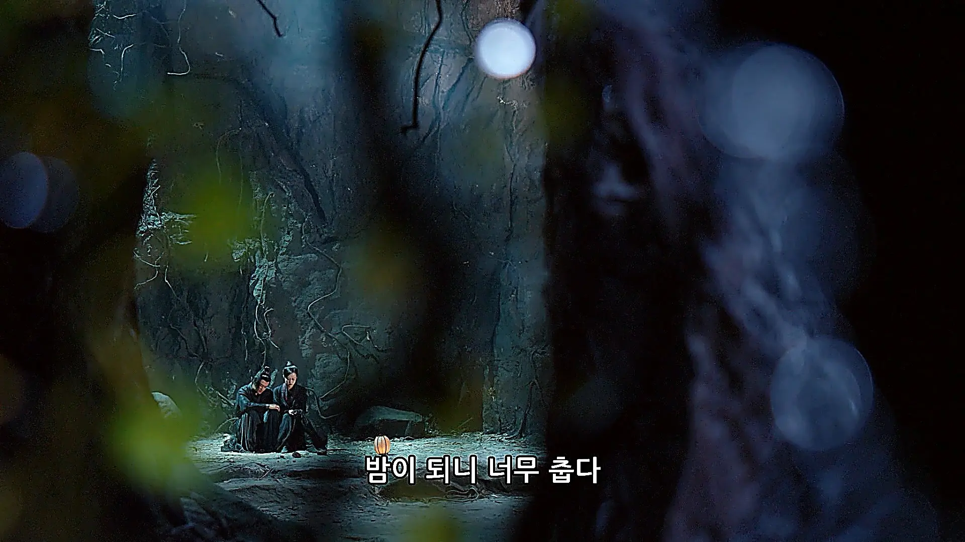 Wide shot of two figures in dark clothing sitting together at the base of a cold, rocky cavern with a small lantern