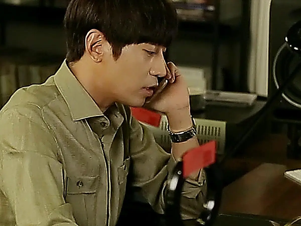 Side profile of a man in an olive button-down shirt holding a phone to his ear at a desk