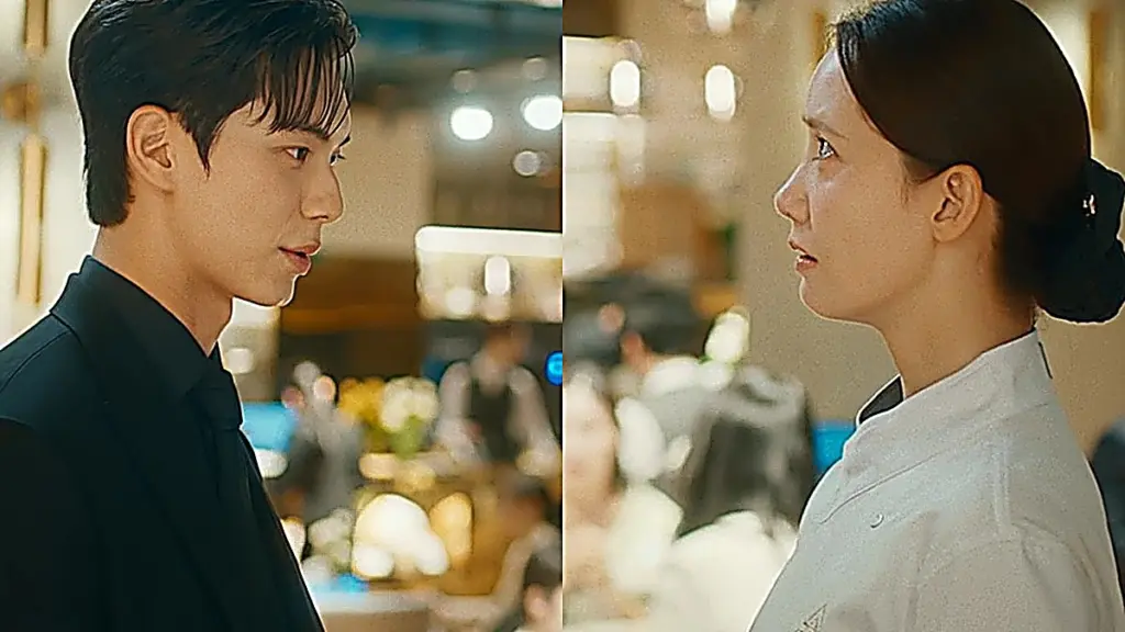 A man in a black modern suit and a woman in a white chef coat facing each other in profile inside a restaurant