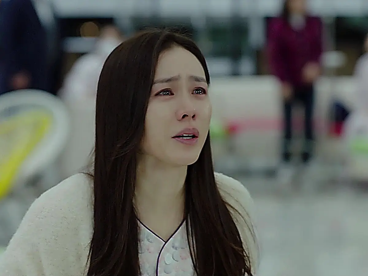 A tearful woman wearing a soft cream knit cardigan over hospital attire.