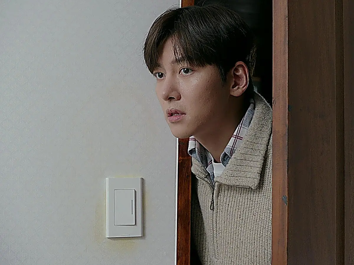 A young man in a beige quarter-zip ribbed knit sweater peering into a room through an open doorway