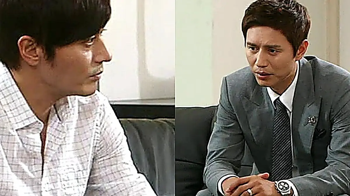 Two men seated across from each other in an indoor setting, one in a white collared shirt and the other in a grey suit and patterned tie